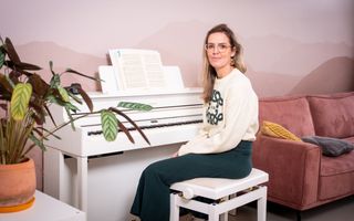 Jonge vrouw in witte trui en blauwe rok zit achter een witte piano. Rechts van haar een roze bank, links een plant.