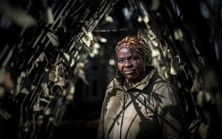 Zwarte vrouw in groene jas met een hoofddoek van bruine stof met blaadjesmotief kijkt glimlachend in de cameralens, vanachter een haag van bladeren.