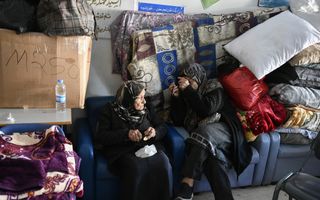 Twee gesluierde vrouwen zitten op bank, naast stapel dekens en kussens.
