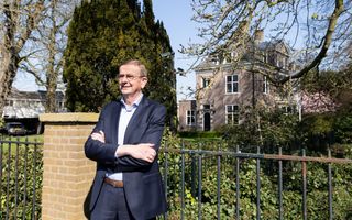 Man van middelbare leeftijd in donkerblauw pak staat voor een hek met op de achtergrond struiken en een statig pand.