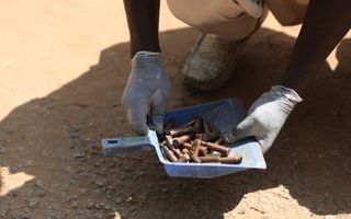 Op de bruine grond zit iemand gehurkt. Hij houdt in gehandschoende handen een blauw bakje met koperen kogelhulzen vast.