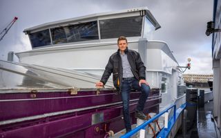 Man in spijkerbroek en werkschoenen staat in het gangboord van een binnenvaartschip en leunt met zijn ene hand op de luiken van het ruim. 