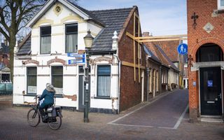 Scheef witgepleisterd huis naast steeg met verstevigingsbalken. Op de voorgrond fietst een vrouw.