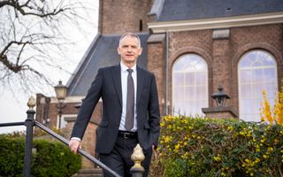 Man in een donker pak met een stropdas staat voor een kerkgebouw met grote ramen.