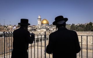Twee in het zwart geklede mannen met zwarte hoeden staan voor een zwart hek. Daarachter ligt de oude stad van Jeruzalem.