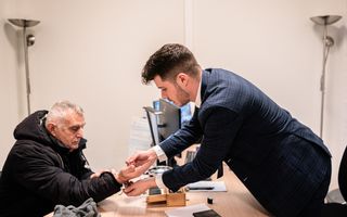 Een man in een donkere jas laat gouden voorwerpen zien aan een jongere man in een blauw pak.