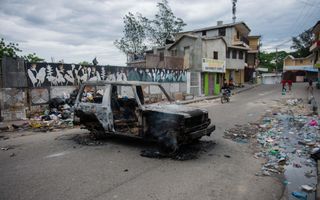 Achter een nog rokend karkas van een auto lopen mensen op een straat waar langs de kanten veel huisvuil ligt opgestapeld. Geelgekleurde huizen staan op de achtergrond. Het is bewolkt. 