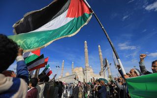 Donkergekleurde mensen met tulbanden en andere hoofddeksels op zwaaien met Palestijnse en Iraanse vlaggen voor een moskee met hoge minaretten en koepels. De lucht is stralend blauw.