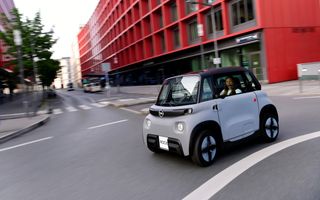 Een grijze brommobiel van het merk Opel rijdt door een stedelijke omgeving met een roodgekleurd gebouw op de achtergrond. 
