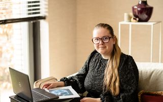 Jonge vrouw met bril en zwarte jurk kijkt in de camera. Ze zit op een bank achter een laptop en een uitgeprint werkstuk.