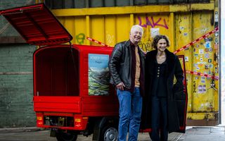 Een man en een vrouw staan voor een klein rood autootje. Op de achtergrond is een gele container te zien.