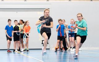 Twee meisjes rennen fanatiek in een gymzaal. Het ene meisje heeft een blauwe band in haar handen en het andere meisje een rode. Op de achtergrond twee teams.