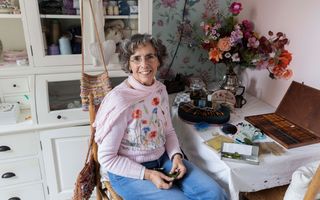 Een vrouw in roze trui met een fleurig veldboeket erop zit bij allerlei handwerkspullen die ze gebruikt voor haar priegelwerk.