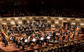 In een grote concertzaal zit een orkest te spelen. Op de bok staat een dirigent.