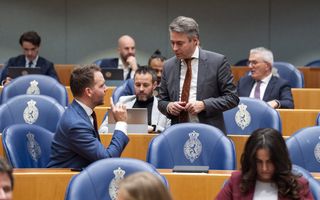 Ongeveer tien keurig geklede mannen en vrouwen zitten op blauwe stoelen waarop het logo van het Rijk der Nederlanden staat, met elkaar te praten