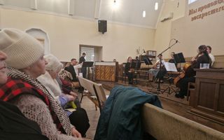Vrouwen zitten in bank. Voor hen op het podium zit een orkest. 