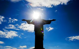 Een beeld van een zegende Christus, daarachter een felle zon, blauwe lucht en enkele wolken.