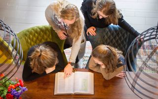 Vier meisjes staan bij tafel, een van hen wijst in openliggende Bijbel iets aan.