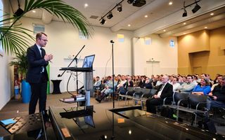 Een jonge man, bril, haar in een scheiding, spreekt van achter een katheder een zaal vol mensen toe.