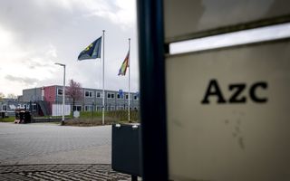 Op de voorgrond een bord waarop ”AZC” staat, op de achtergrond een woningencomplex met enkele vlaggenmasten ervoor.
