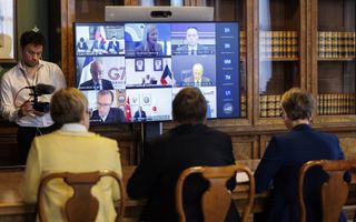Drie mensen zitten achter een tafel voor een groot beeldscherm. Daarachter een boekenkast gevuld met oude boeken. 