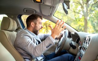 Een man met kort bruin haar, grijze colbert en nette broek zit in een auto en schreeuwt terwijl hij gefrustreerde handgebaren maakt.