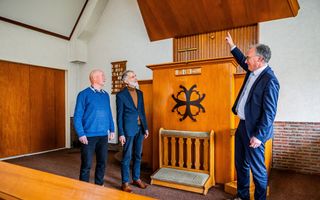 Drie mannen, in het blauw gekleed, staan bij de kansel van de kerk. De man links, in pak, wijst met zijn vinger naar boven.