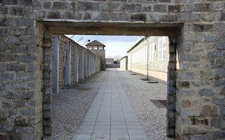 Doorkijkje door een poort in een muur. Erachter een gebouw rechts en een muur met prikkeldraad en wachttoren links.