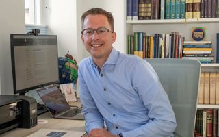 Relatief jonge man met moderne bril en in een blauw overhemd zit achter een bureau met een computerscherm.