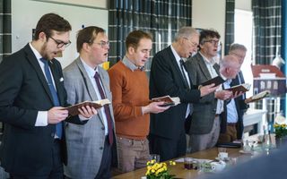 Zeven mannen staan naast elkaar achter een tafel te zingen, met een psalmboek in hun handen.