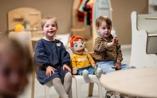 Twee kinderen zitten op een stoeltje. Tussen hen in zit een pop met een gele trui waar de naam Bas op staat. 