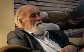 oude man met wit haar en witte baard in grijs jasje over een wit overhemd zit op een bank en kijkt droevig.