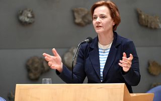 Glimlachende vrouw van middelbare leeftijd en kort rood haar in donkerblauwe colbert staat achter een katheder en maakt wijds handgebaar.