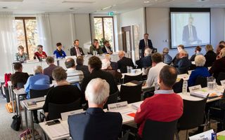 Een vergaderzaal vol mensen achter lange tafels.