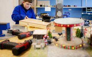 Een leerling in een blauwe overall is bezig met het in elkaar zetten van een houten laatje. Hij kijkt ingespannen en geconcentreerd. Op het werkblad liggen twee boren.