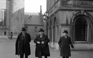 Zwart-witbeeld van drie mannen in een donker pak met een lange jas en een hoed lopend over een plein tussen historische gebouwen.