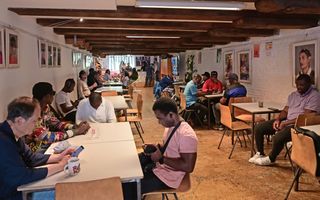 Mannen zitten in een zaal aan verschillende tafels. Aan de wanden hangen schilderijen. 