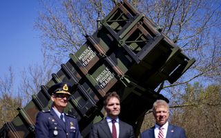 Drie mannen, een in militair uniform, staan voor een groot rechthoekig apparaat in legerkleuren, waar Missile Round Trainer op staat.