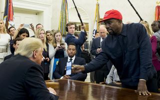 Een zwarte man met een rode pet op en een donkerblauwe blouse aan staat voor een groot bureau waarachter een man met blond haar in pak zit. Op de achtergrond staan allemaal mannen en vrouwen in nette kleding. 