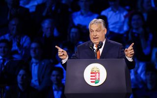 Man met grijs haar en oranje das aan het spreken voor twee microfoons en met publiek achter zich.