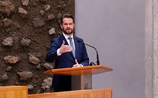 Man van middelbare leeftijd met baard in donkerblauw pak achter spreekkatheder.