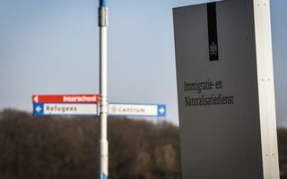 Wit bord met daarop in blauwe letters de tekst ”Immigratie- en Naturalisatiedienst” met op de achtergrond wegwijzers voor vluchtelingen.