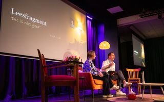 Een man en vrouw zitten voor een groot scherm op een bank. Achter hen een brandende lamp. 