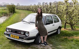 Jonge vrouw zit op de motorkap van een witte Golf GTI. De auto staat in een wijngaard met bloeiende fruitbomen.