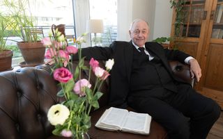 Een man in een zwart pak zit op een bank. Naast hem ligt een opengeslagen Bijbel. Op de voorgrond staat een boeket bloemen.