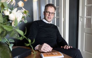 Een man met donker haar, een bril en een wit overhemd met daarover een zwarte trui zit aan een tafel.