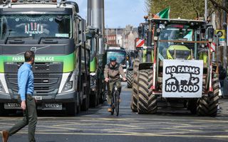 Vrachtwagens en tractoren staan naast en achter elkaar op een straat in een stad. Een fietser en een wandelaar passeren de blokkade. Op een tractor hangt een bord met de tekst: ”No farms, no food”.