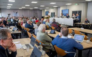 Mannen en vrouwen achter vergadertafels. Voorin, op een podium, een tafel met daarachter vijf personen. 