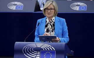 Een dame met blond haar, bril en lichtblauw jasje staat achter een katheder met het embleem van de Europese Unie: een blauw vlak met twaalf gele sterren in een cirkel.