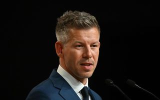 Een 45-jarige man, grijze kuif, blauw pak, tegen een zwarte achtergrond.